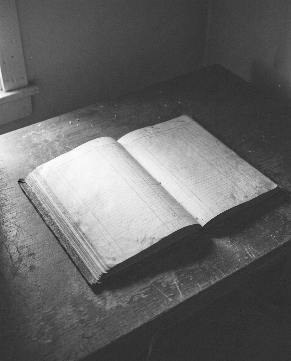 An open bound ledger with faint ruled columns, lying on a dark desk under a single hard light.