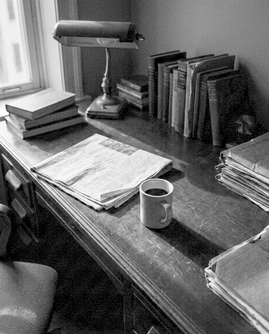 A folded newspaper beside a cooling cup of black coffee on a dark wooden desk in a private office.